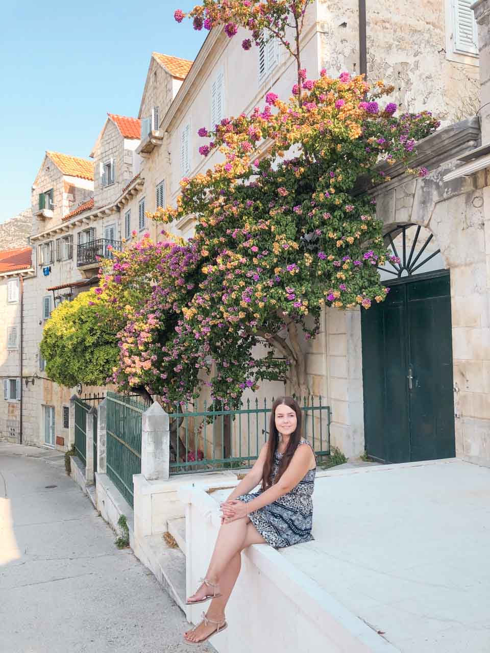 A girl sitting on top of a white stone wall in Bol, Croatia with a purple tree in the background