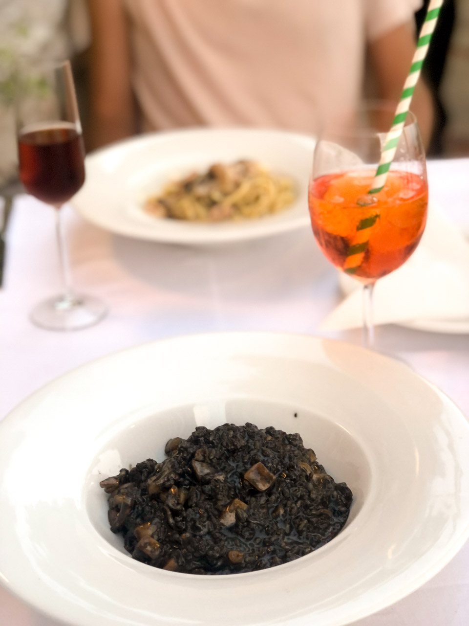 Plates of black risotto and truffle pasta on a table next to an Aperol Spritz and a glass of Prošek wine