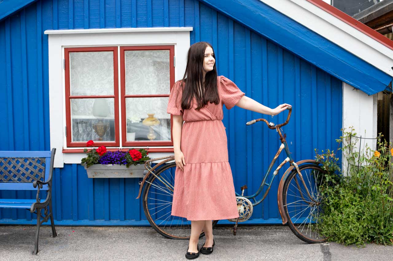 A brunette in a pink dress holding a bike in front of a blue wooden house