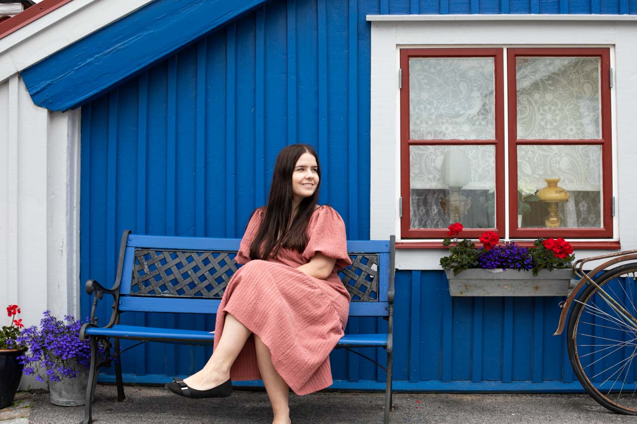 A smiling brunette in a pink dress sitting on a blue bench outside a blue wooden house