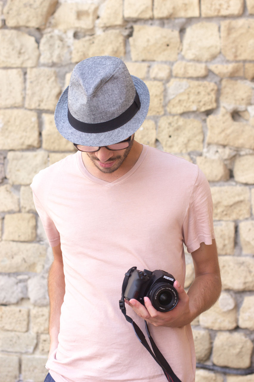 A man in a hat looking down and holding a camera, standing in front of a brick wall in Naples, Italy