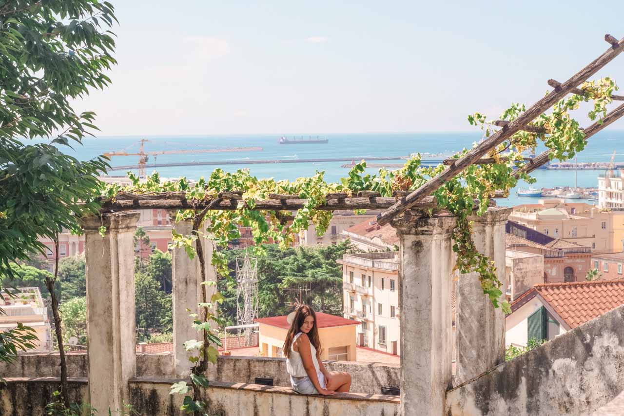Girl in a white crop top and denim shorts sitting on the ledge of Giardino della Minerva in Salerno