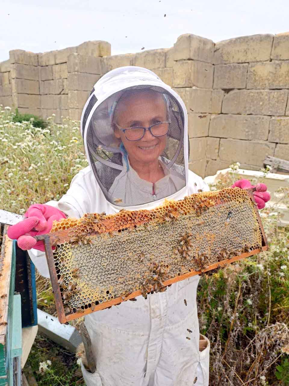 A beekeeper smiling at the camera as she is holding a hive frame with capped honey cells