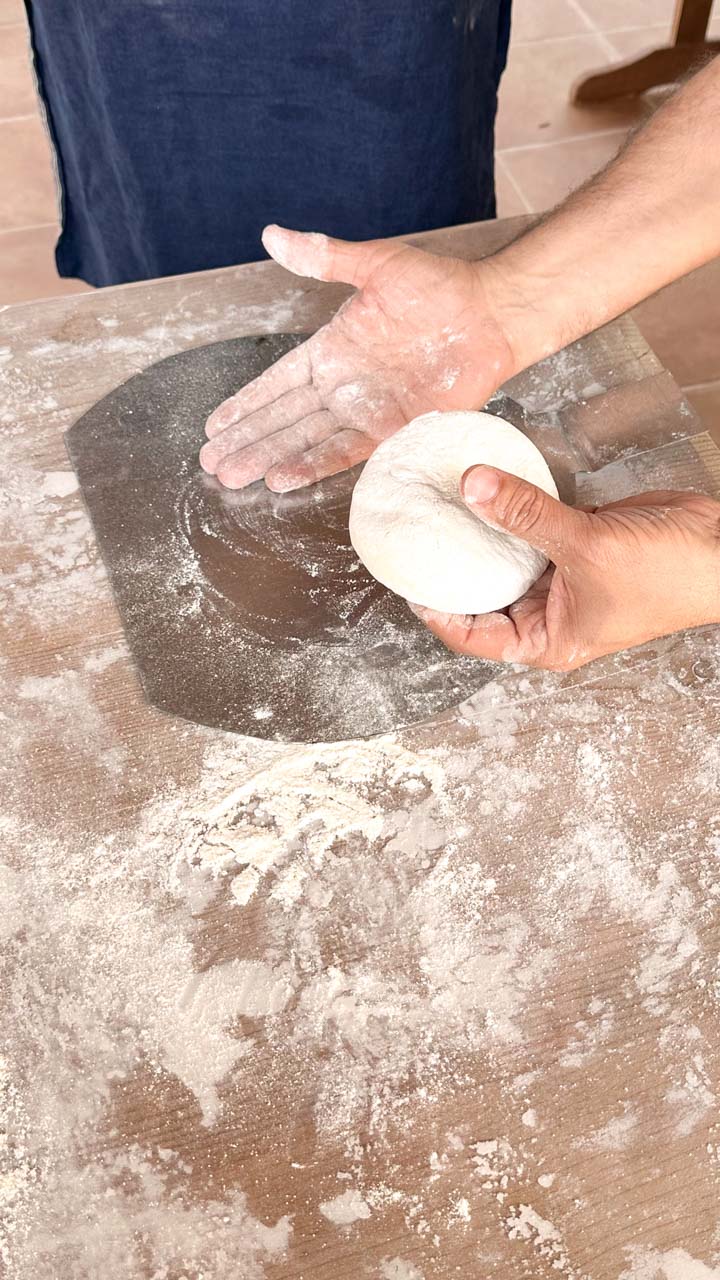 A man holding a ball of pizza dough in one hand as he's pointing to a pizza peel with his other hand