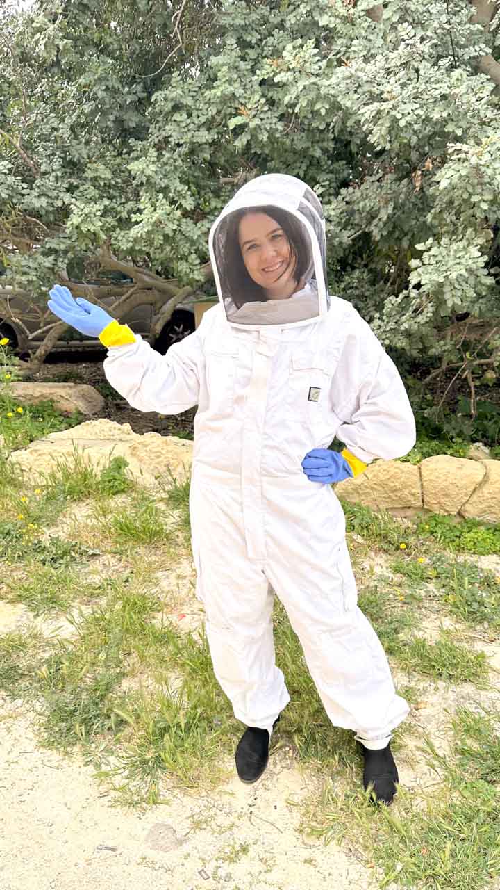 A smiling woman in a beekeeper's suit happily pointing at the tree behind her