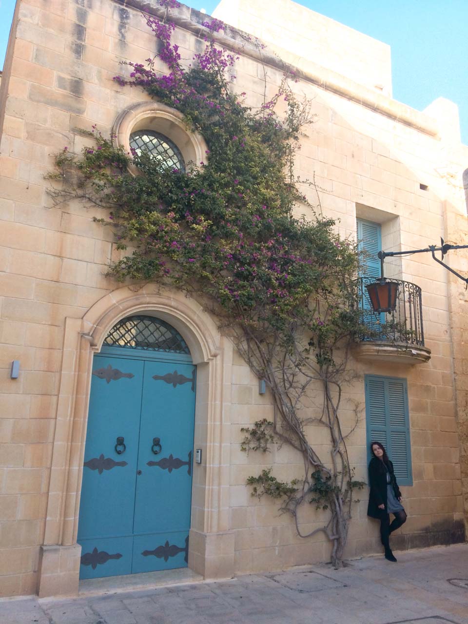 Girl standing next to a doorway and a wall covered with blooming flowers and strands of ivy in Mdina