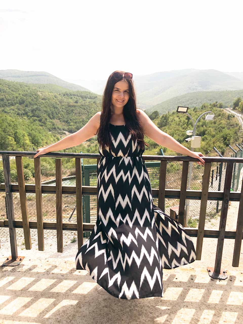 Girl in a maxi dress smiling at the camera and she's standing on a viewing terrace at the Pristina Bear Sanctuary