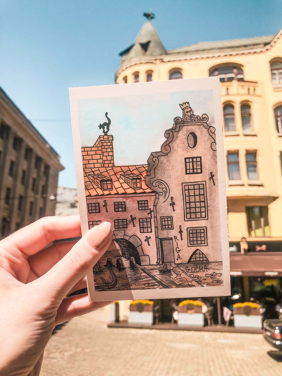 A woman's hand holding a postcard with Riga's Cat House on it in front of the actual Cat House
