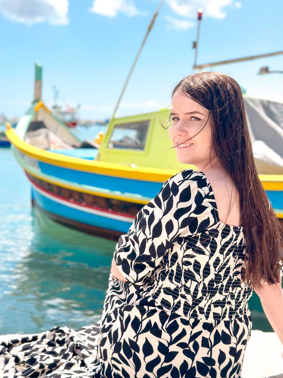 A brunette in a black and white dress smiling at the camera with a traditional Maltese fishing boat, named luzzu, behind her