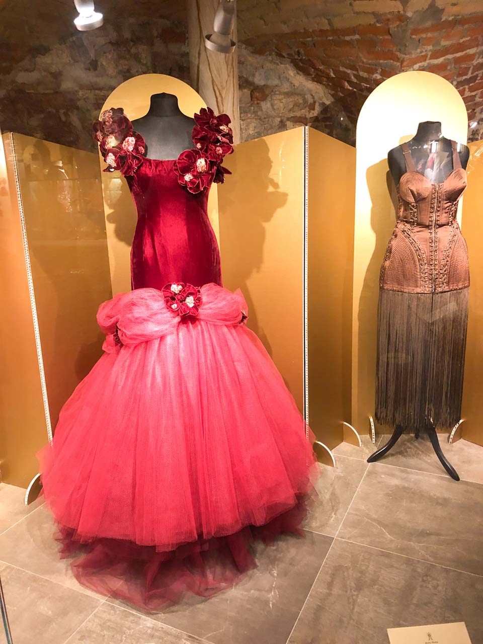 A red gown with faux flowers and a brown corset dress with tassels on display at the Fashion Museum in Riga