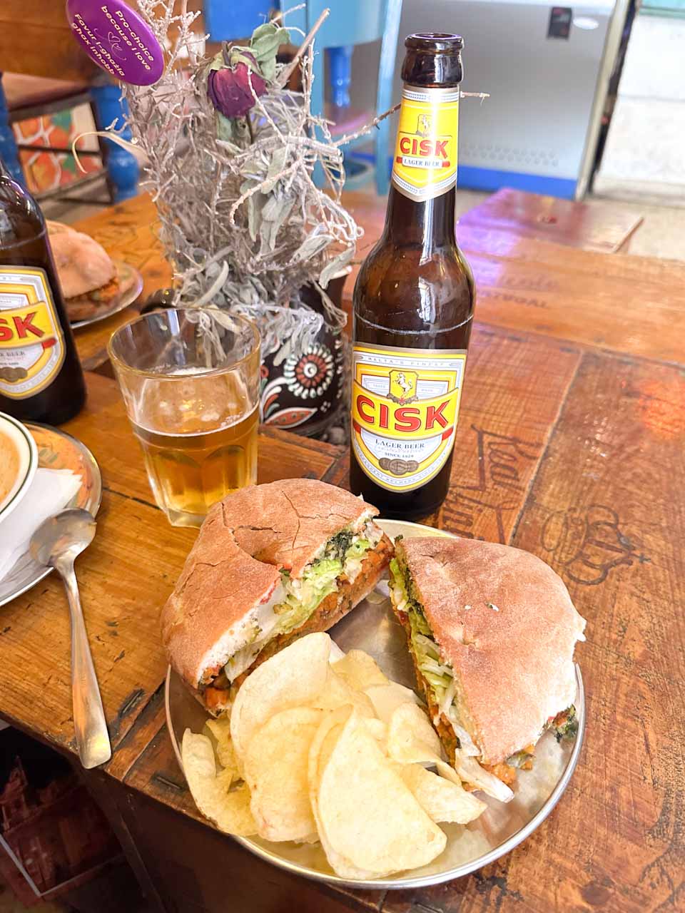 A ftira with a side of crisps, a bottle of Cisk beer, and a glass on a wooden table
