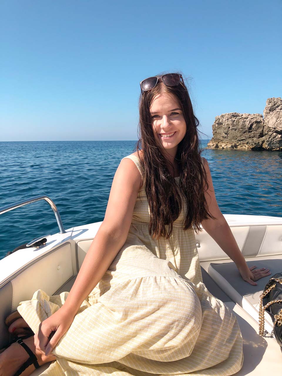 Brunette in a white and yellow maxi dress smiling as she is sitting on a boat deck outside the Blue Cave in Kotor Bay