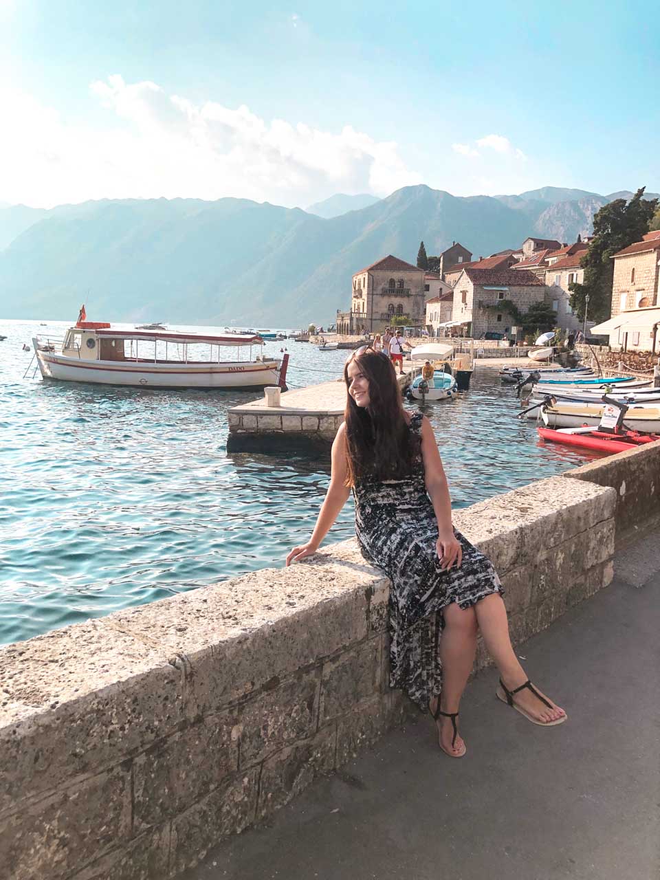 Girl in a black and white dress sitting on a stone wall with the boats in Perast harbour behind her