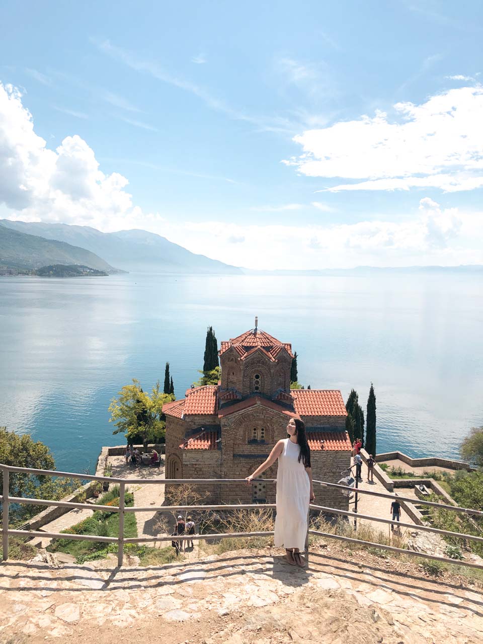 Woman in a white maxi dress standing on a platform overlooking the Church of St. John at Kaneo in Ohrid