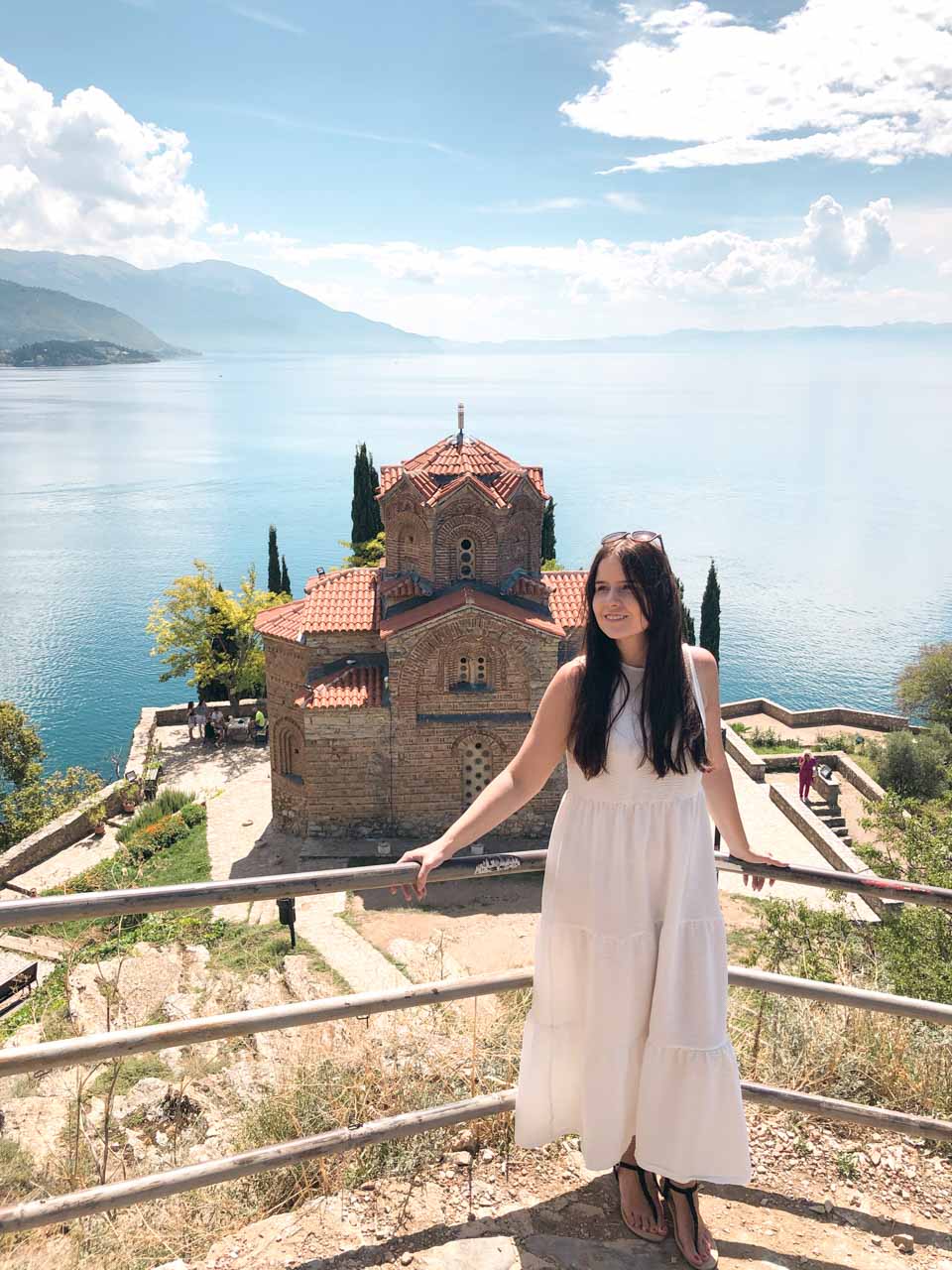 Woman in a white maxi dress standing on a platform overlooking the Church of St. John at Kaneo in Ohrid