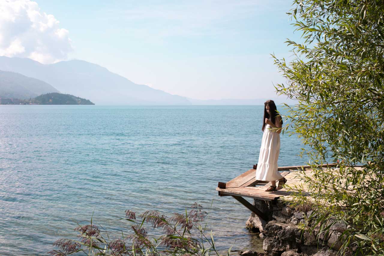 Woman in a white maxi dress standing on the Ohrid Boardwalk looking down at the water