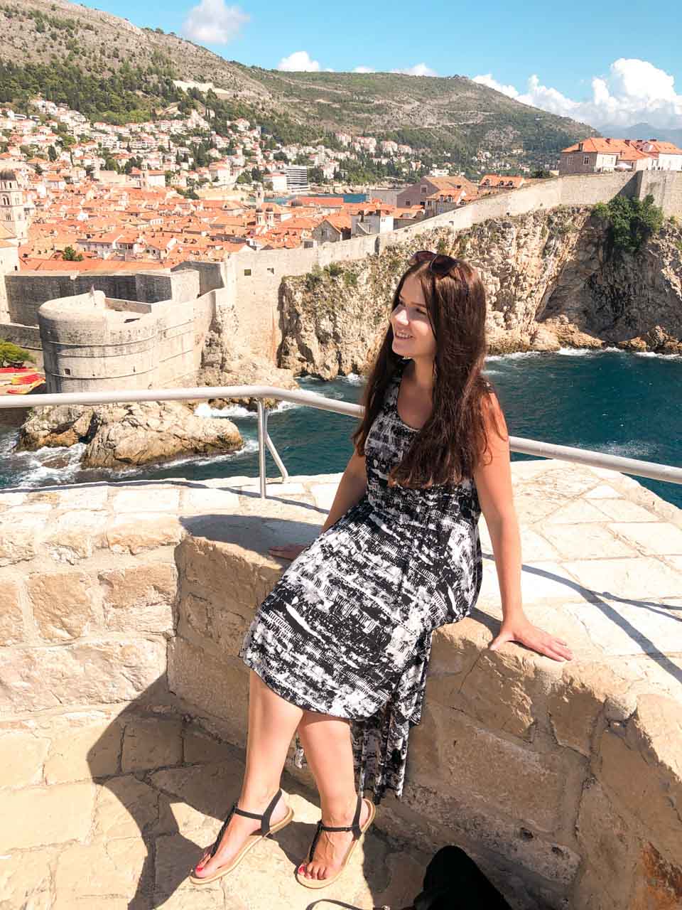 Brunette in a black and white high-low dress sitting on the ledge of the Lovrijenac Fortress in Dubrovnik, Croatia