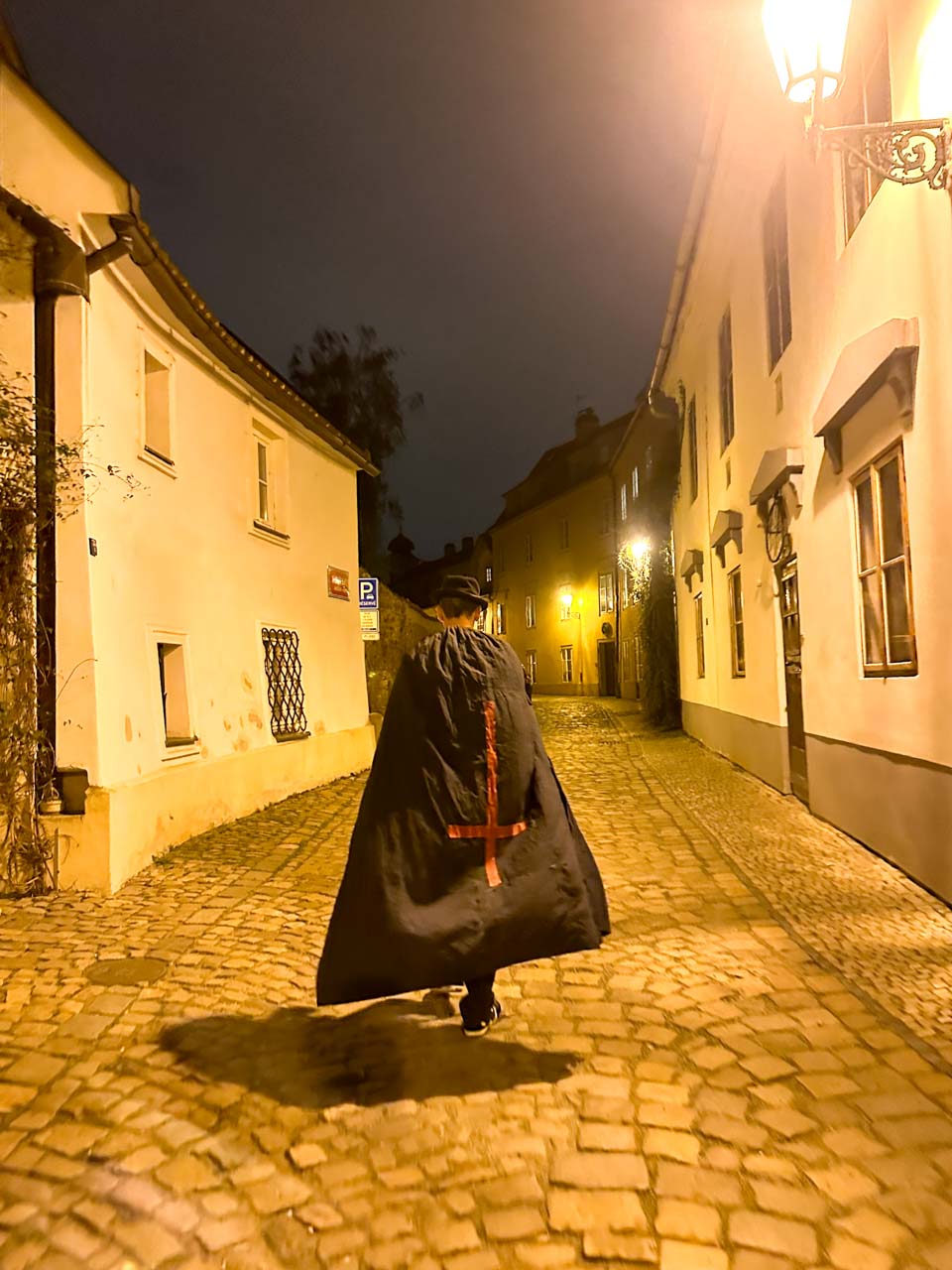 Man in a black cloak with a red cross on it walking down a street in Prague at night