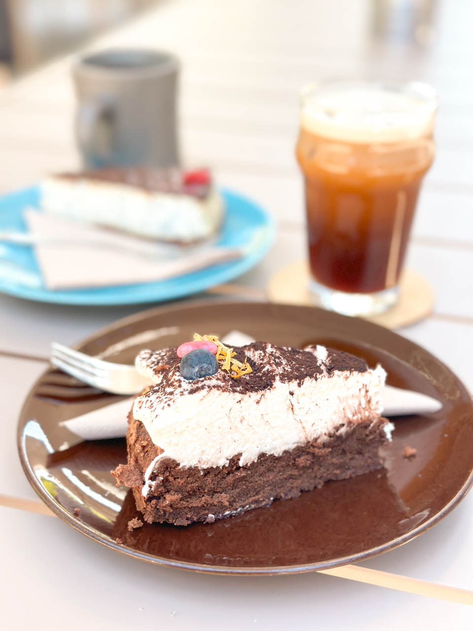 A close-up of a chocolate cake slice with whipped cream, cocoa powder, and a blueberry topping next to a glass of nitro coffee