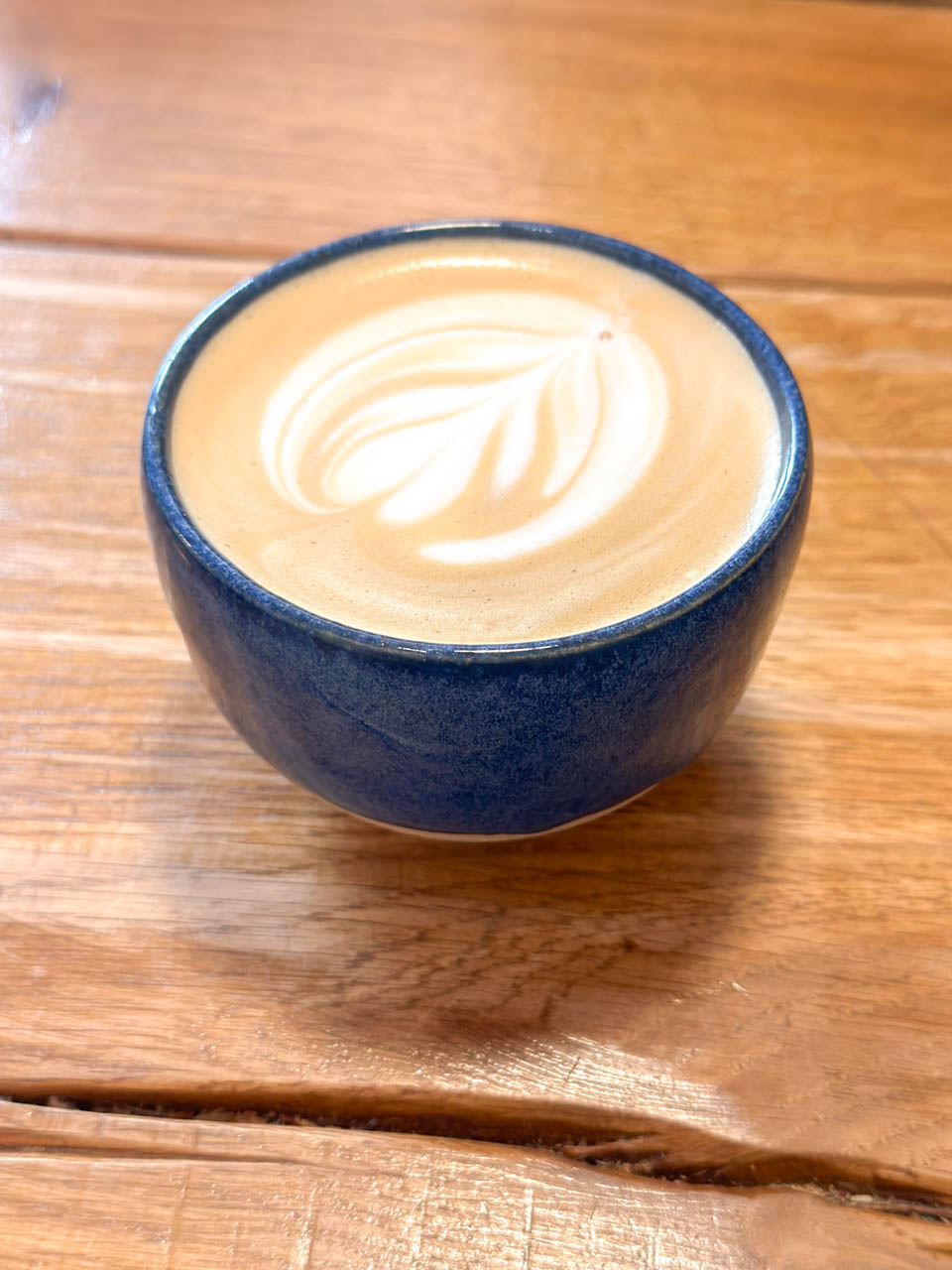 Close-up of a steaming latte with leaf latte art in a blue ceramic mug, placed on a textured wooden table
