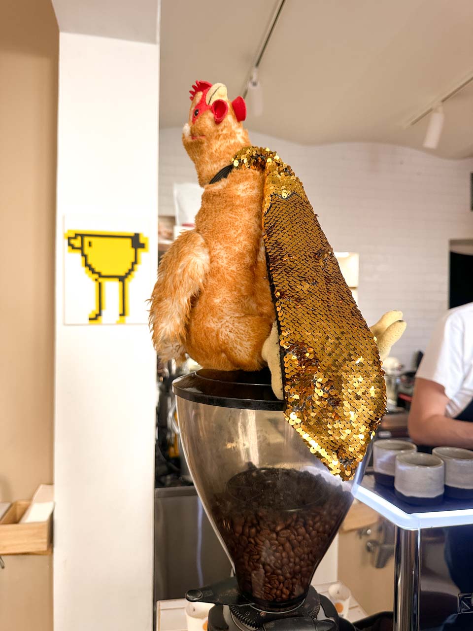 A plush chicken toy wearing a sequinned tie perched on a coffee grinder at Eggo Truck Brno