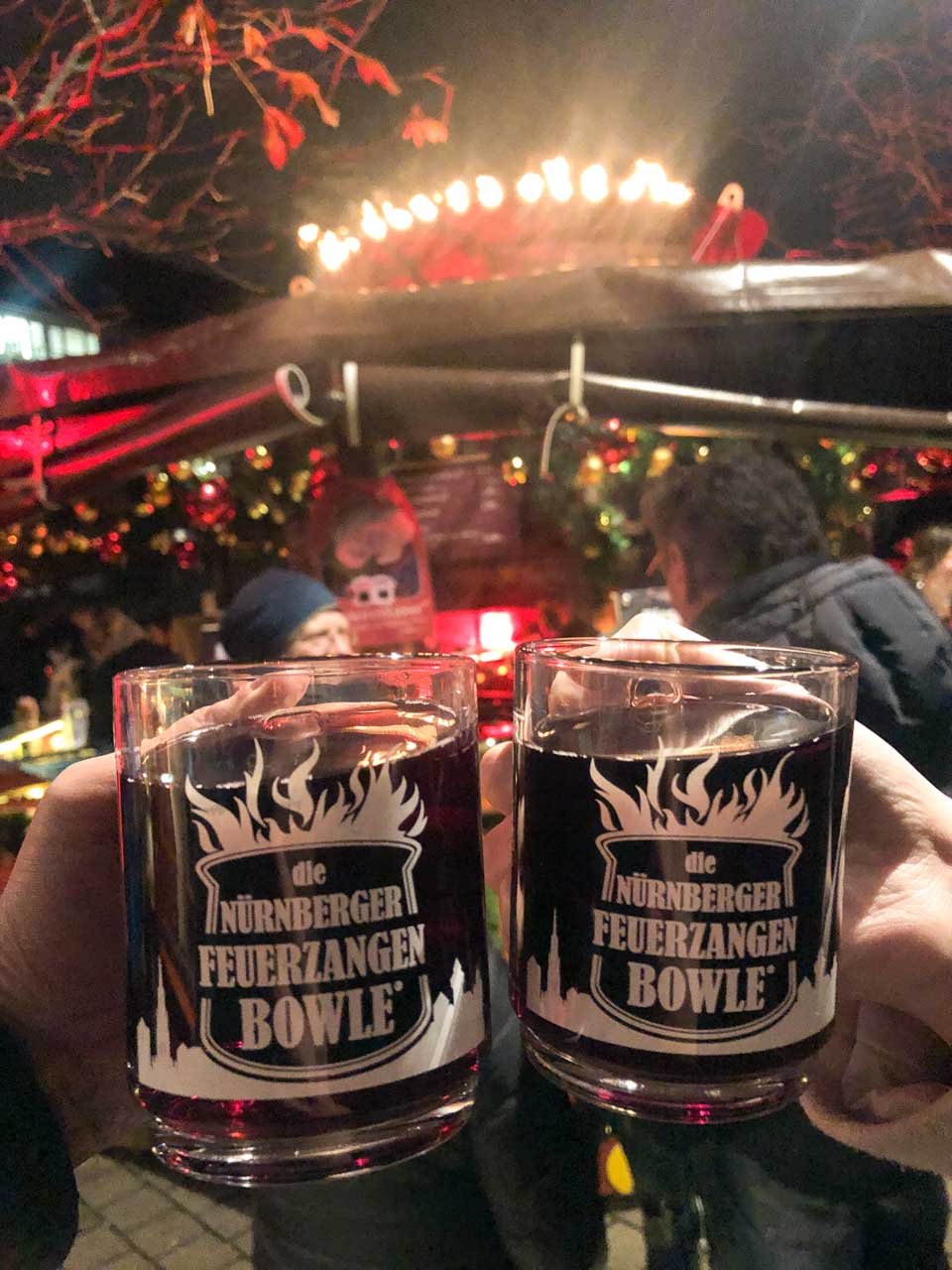 Two hands holding up cups of Nürnberger Feuerzangenbowle at the Nuremberg Christmas Market, with festive lights in the background