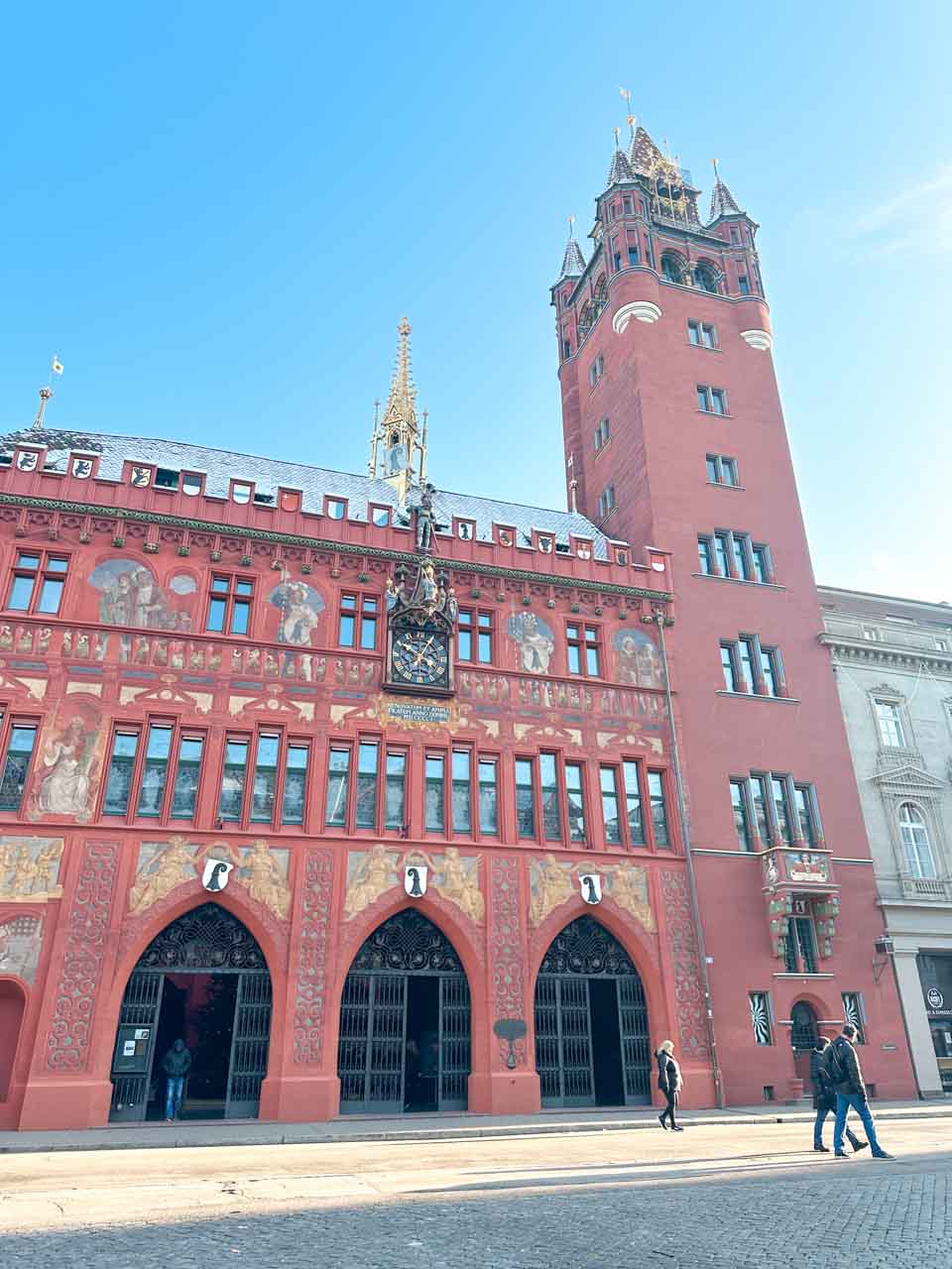 The ornate red facade of the Basel Town Hall with a tower against a clear blue sky