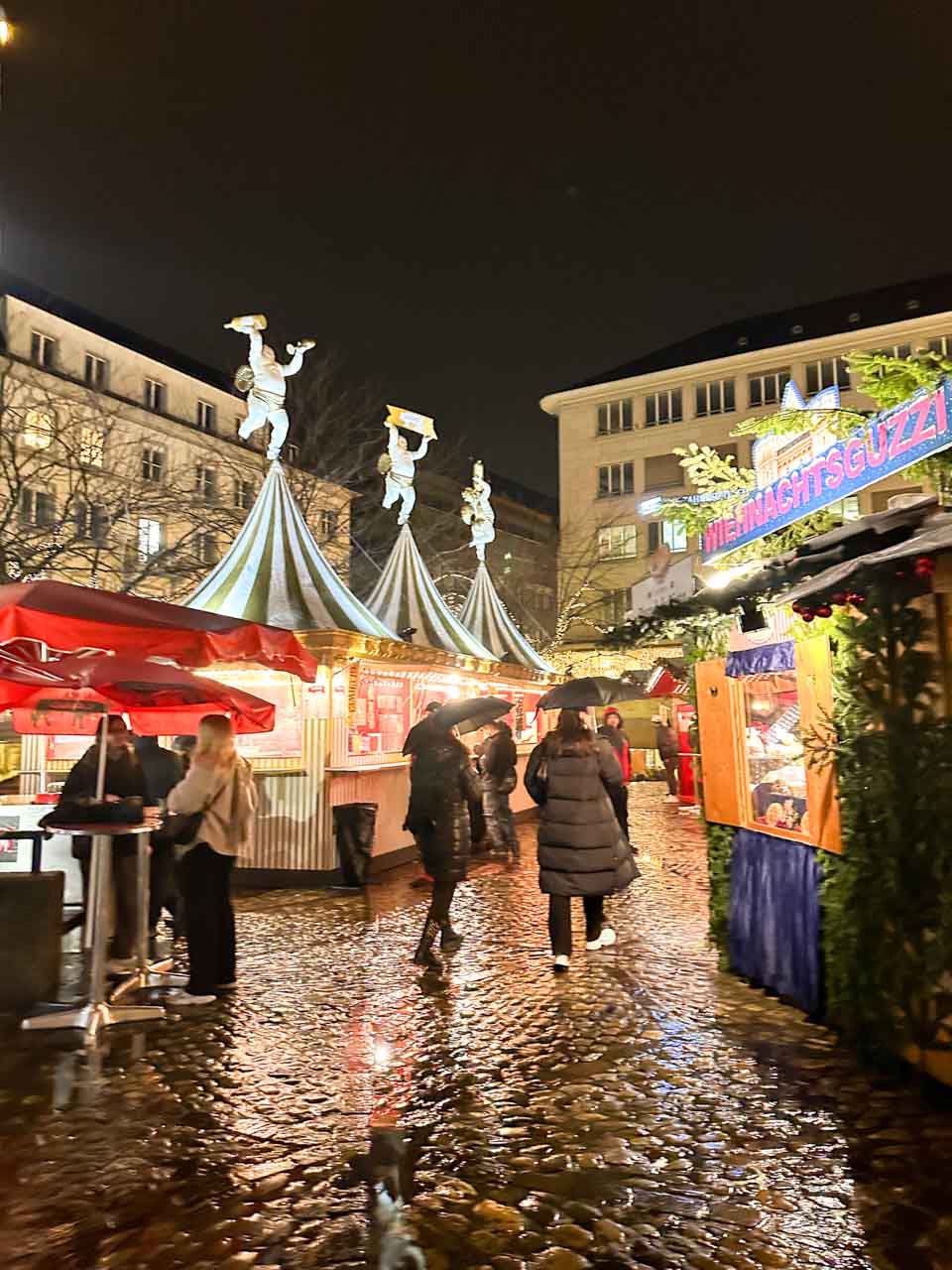 People strolling through the Basel Christmas market on Barfüsserplatz at night, with bright stalls and decorations