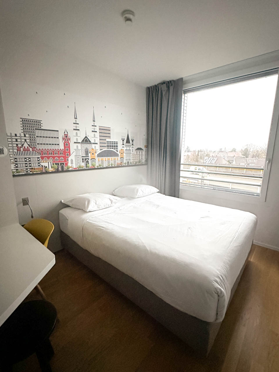 A minimalistic hotel room at B&B Hotel Basel with a large bed and a striking wall mural of Basel's skyline