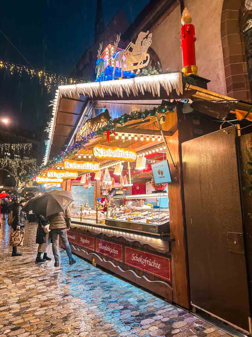 A cosy Christmas market stall at night offering an array of chocolate-covered fruits, with shoppers browsing under a light drizzle