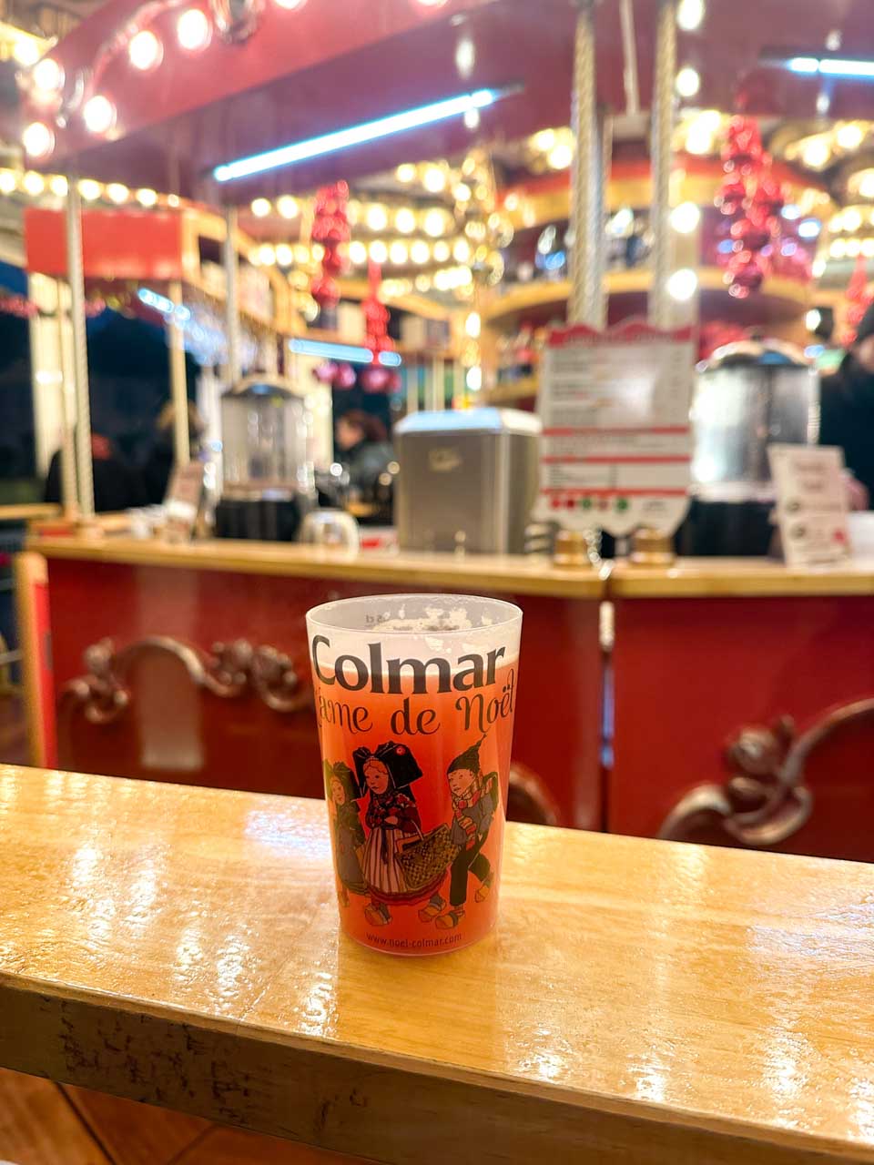 A close-up of a warm cranberry juice cup placed on a wooden counter with a carousel and festive lights softly blurred in the background