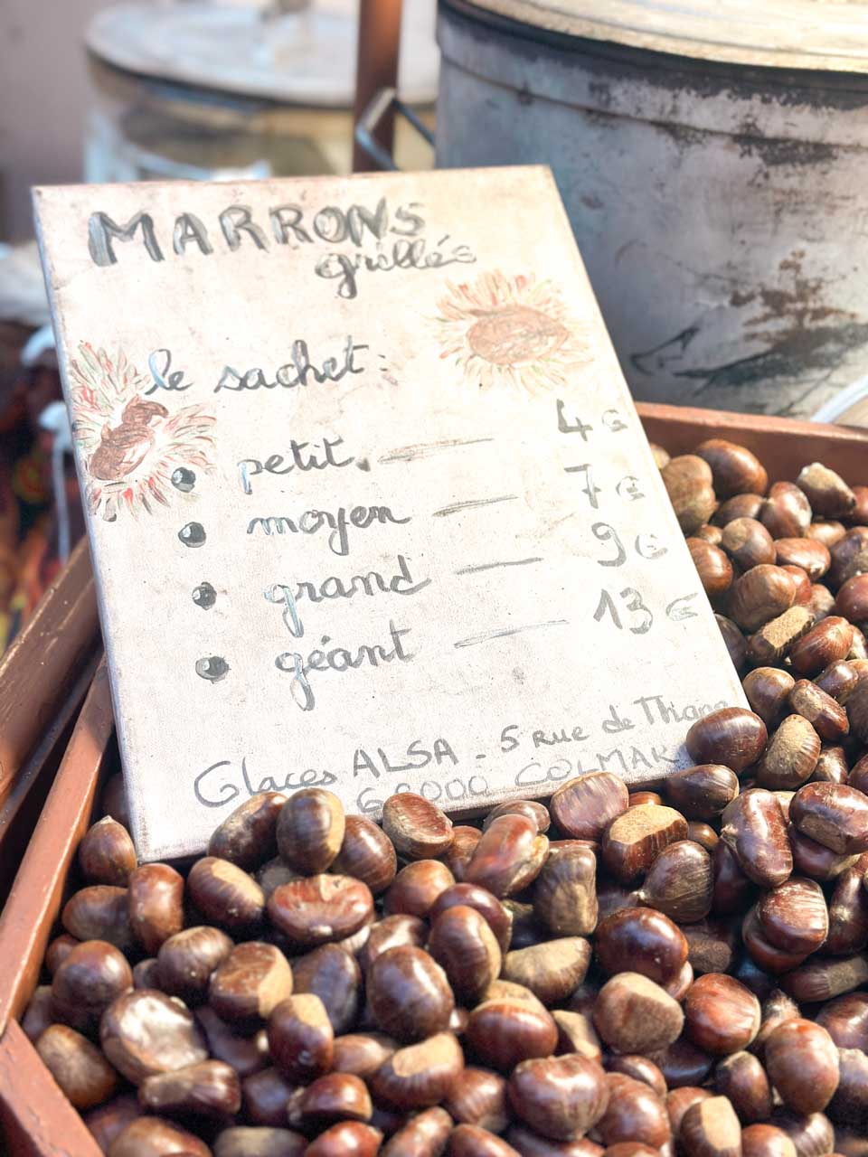 A close-up of roasted chestnuts displayed in a wooden crate, with a hand-painted sign listing prices for different sizes at the Colmar Christmas Markets