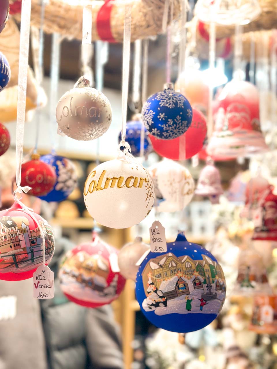 An array of hand-painted Christmas ornaments hanging on display at a market stall, with 'Colmar' and other festive designs