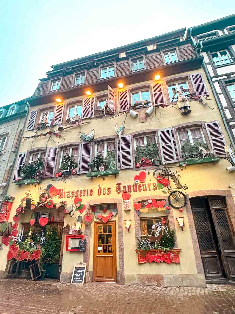 The facade of Brasserie des Tanneurs in Colmar, festively decorated with hearts, hanging greenery, and quirky figurines