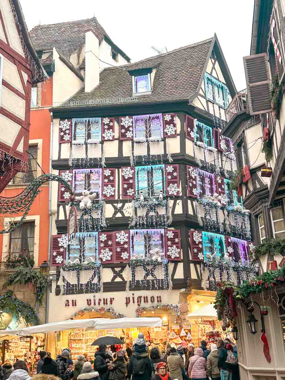 A half-timbered building in Colmar adorned with snowflake decorations and Christmas garlands, with the sign 'Au Vieux Pignon' and a crowd of market-goers in front