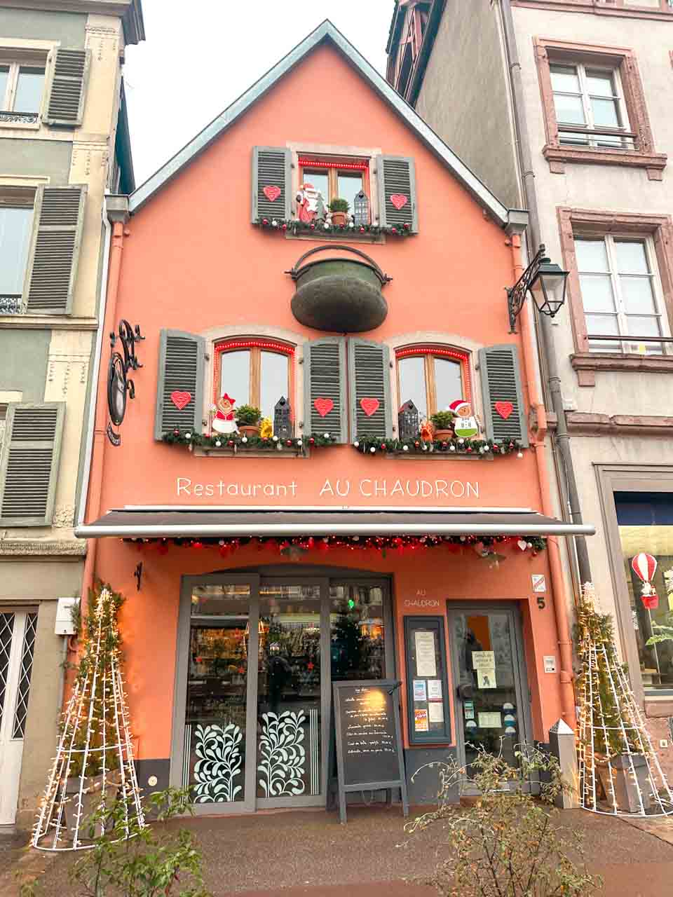 The facade of Restaurant Au Chaudron in Colmar, painted in a vibrant salmon hue with Christmas wreaths and hearts on the shutters, complemented by a minimalist Christmas tree decoration