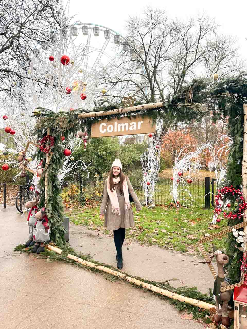 A smiling woman walking through a festive archway labelled 'Colmar', with Christmas decorations and a Ferris wheel in the background