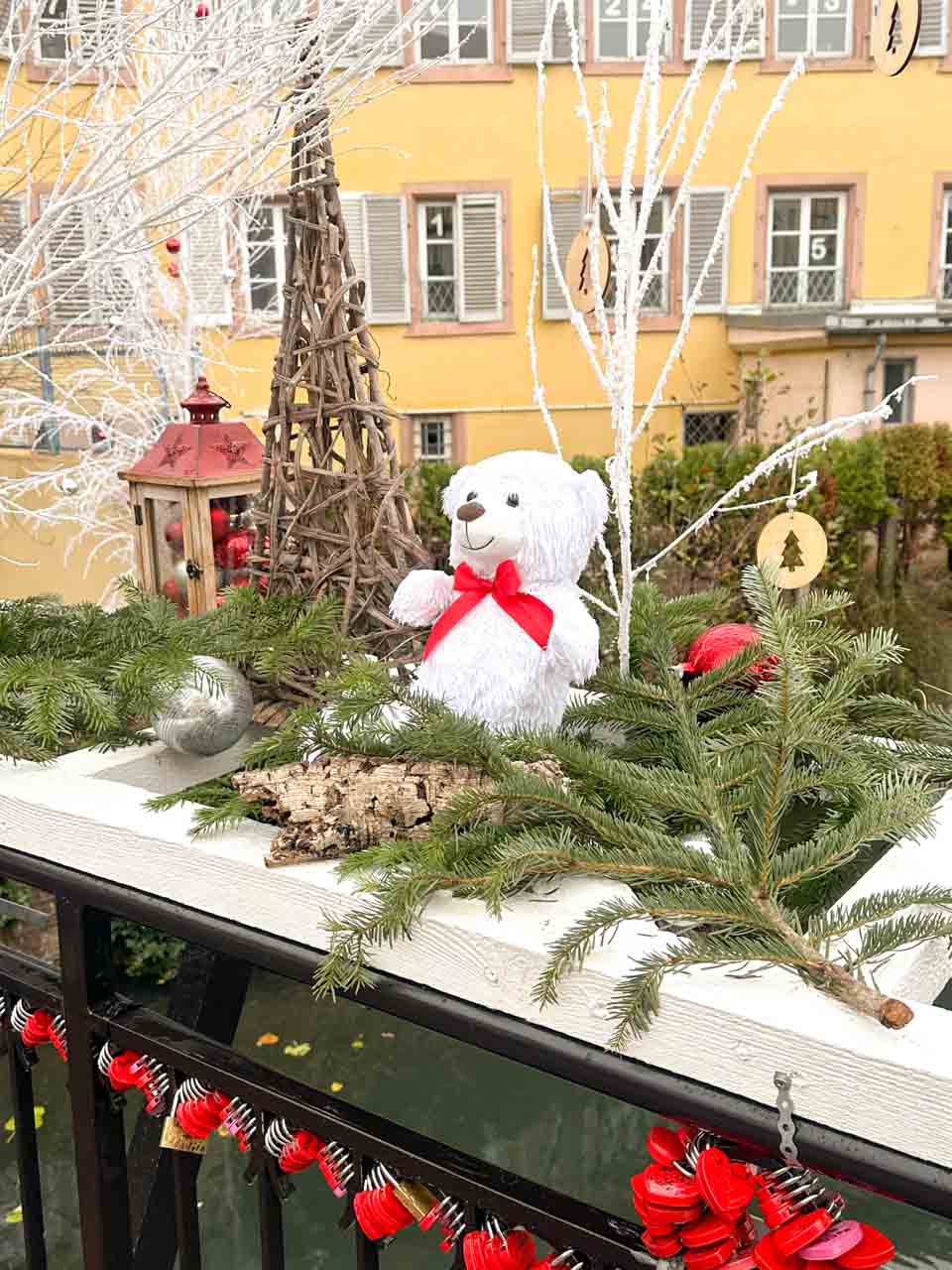 A white teddy bear with a red bow, surrounded by Christmas greenery and ornaments, on a decorated bridge railing in the Petite Venise area of Colmar
