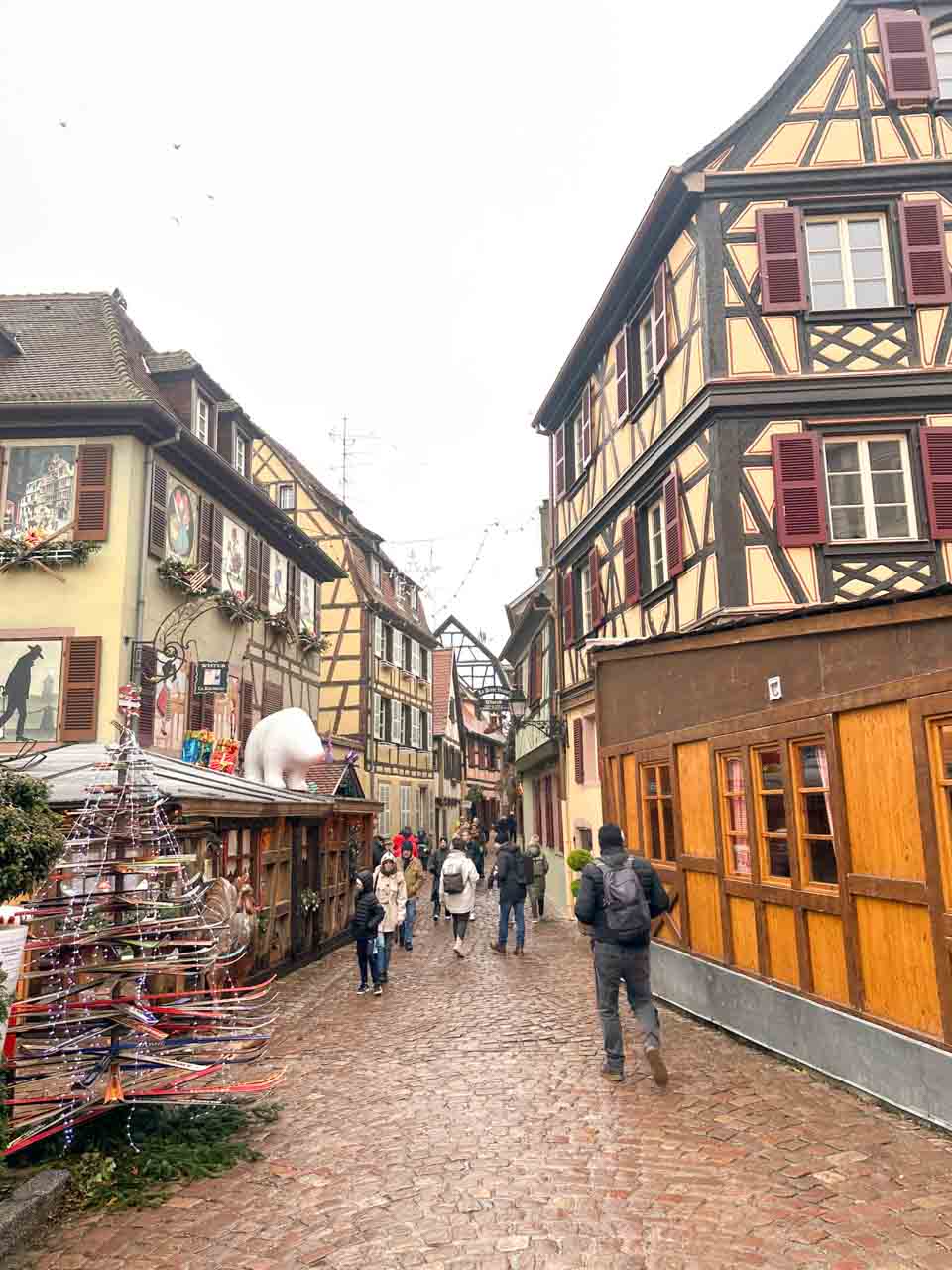 A cobblestone street in Colmar lined with traditional half-timbered houses and a Christmas market stall, with people walking and exploring