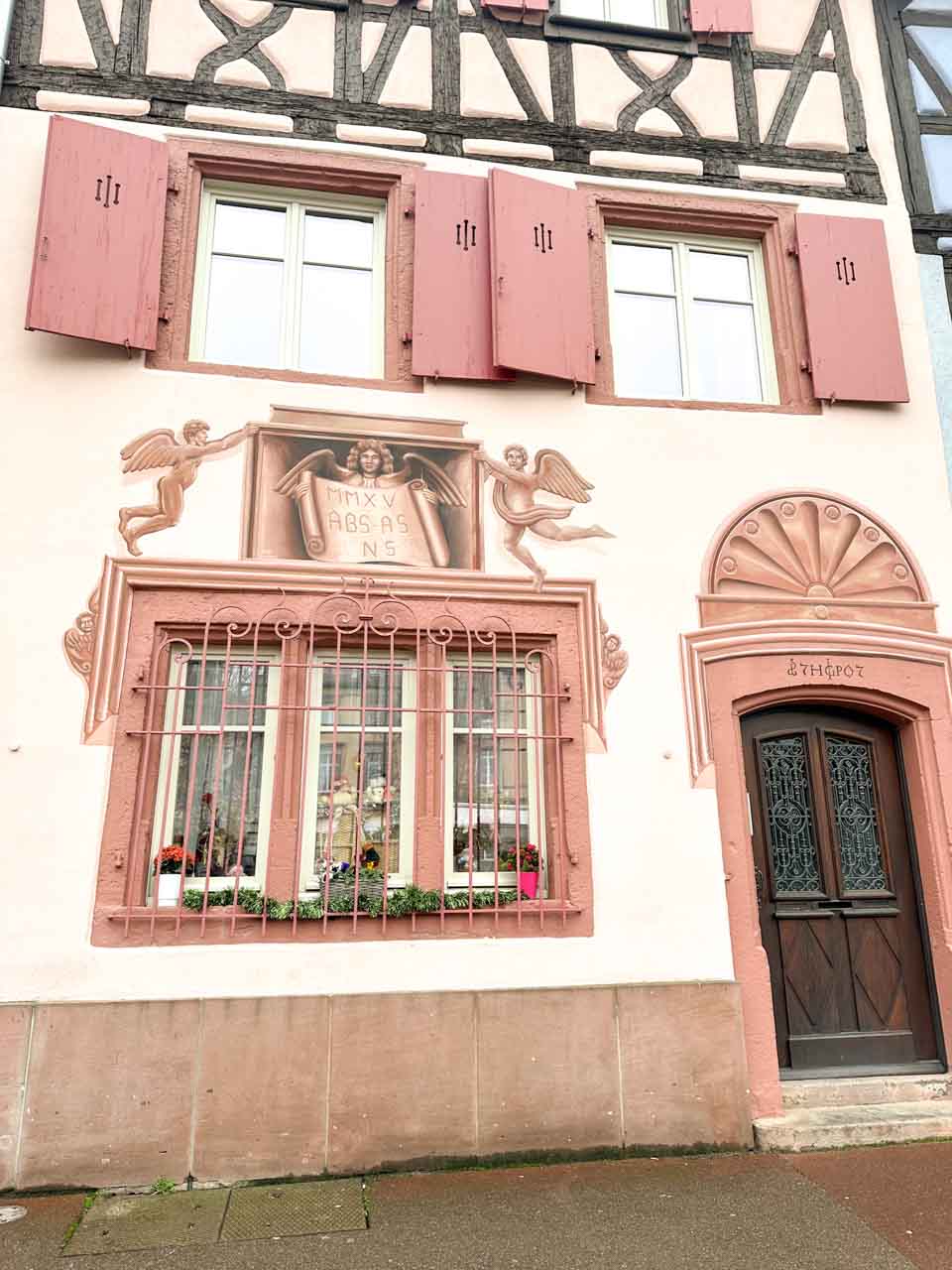 A close-up of a pink traditional Alsatian house with ornate window grills and a fresco between the windows located on Quai de la Poissonnerie in Colmar