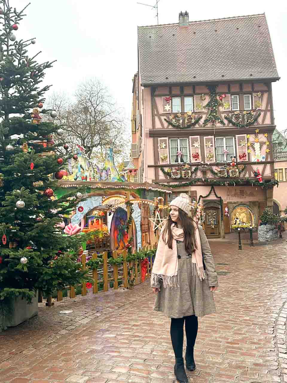 A woman in a warm winter outfit stands in front of a grand Christmas tree and a colourfully decorated half-timbered house in Colmar