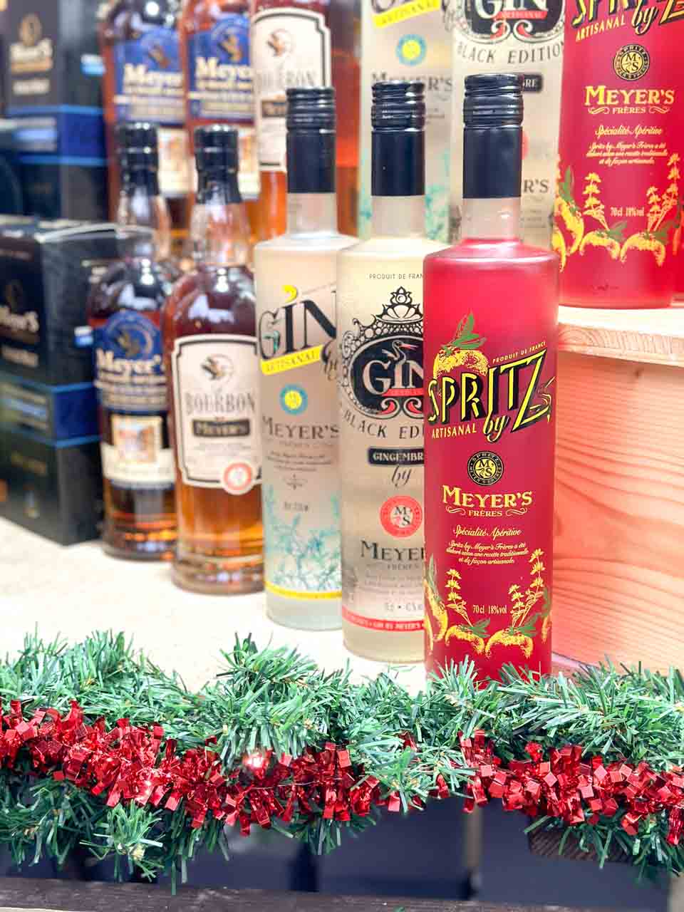 A selection of artisanal spirits and liqueurs, including gin and a bright pink 'Spritz' by Meyer's, displayed among festive garlands at the Colmar Christmas market