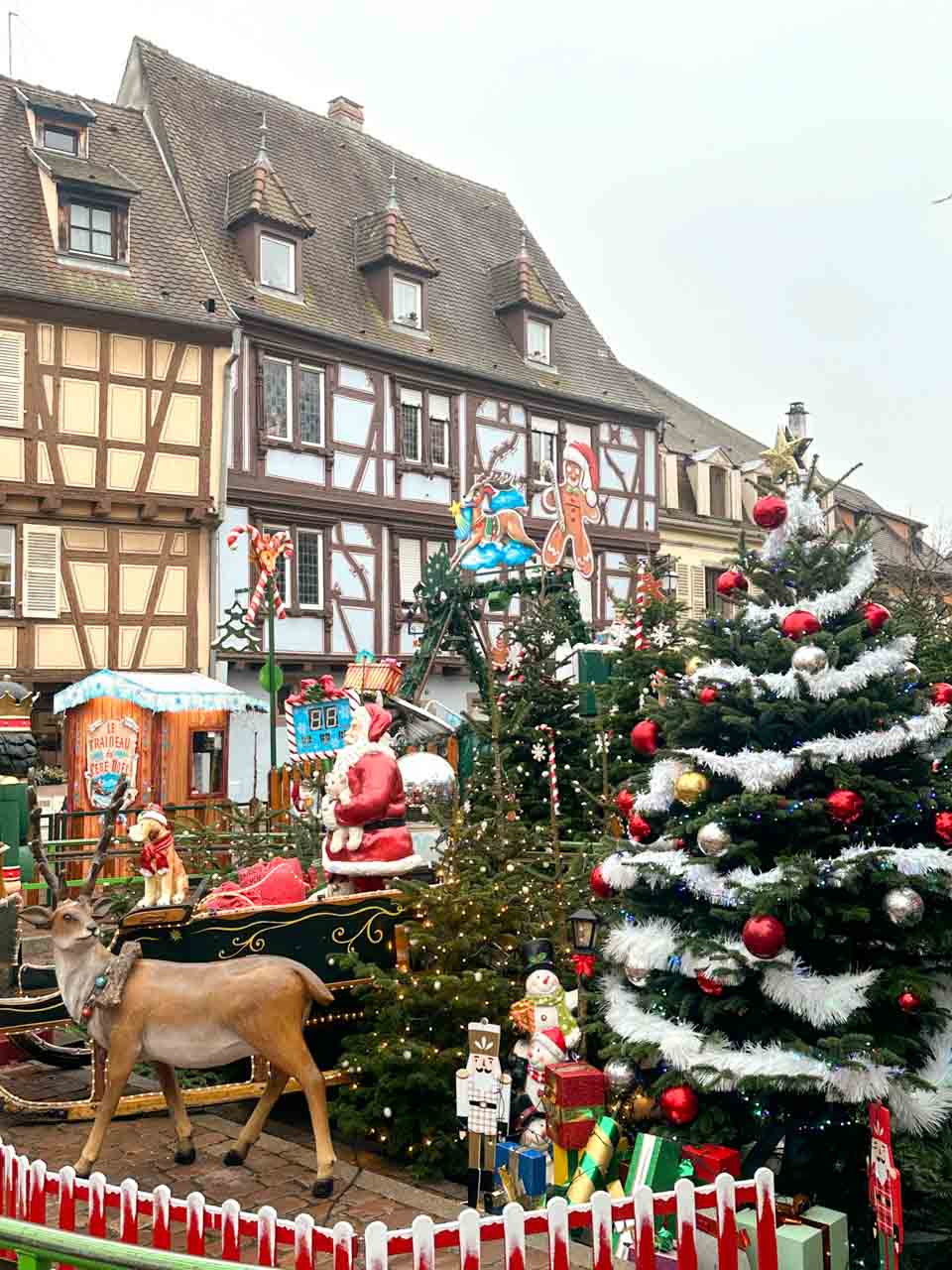 A vibrant Christmas display in Colmar featuring a decorated tree, a life-size Santa in a sleigh, and festive figures, with traditional half-timbered houses in the background