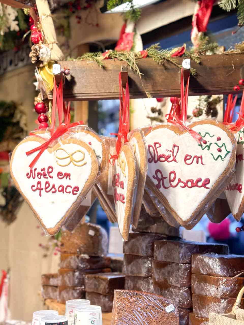 Heart-shaped gingerbread cookies hanging from a market stall, each inscribed with 'No&euml;l en Alsace' (Christmas in Alsace)