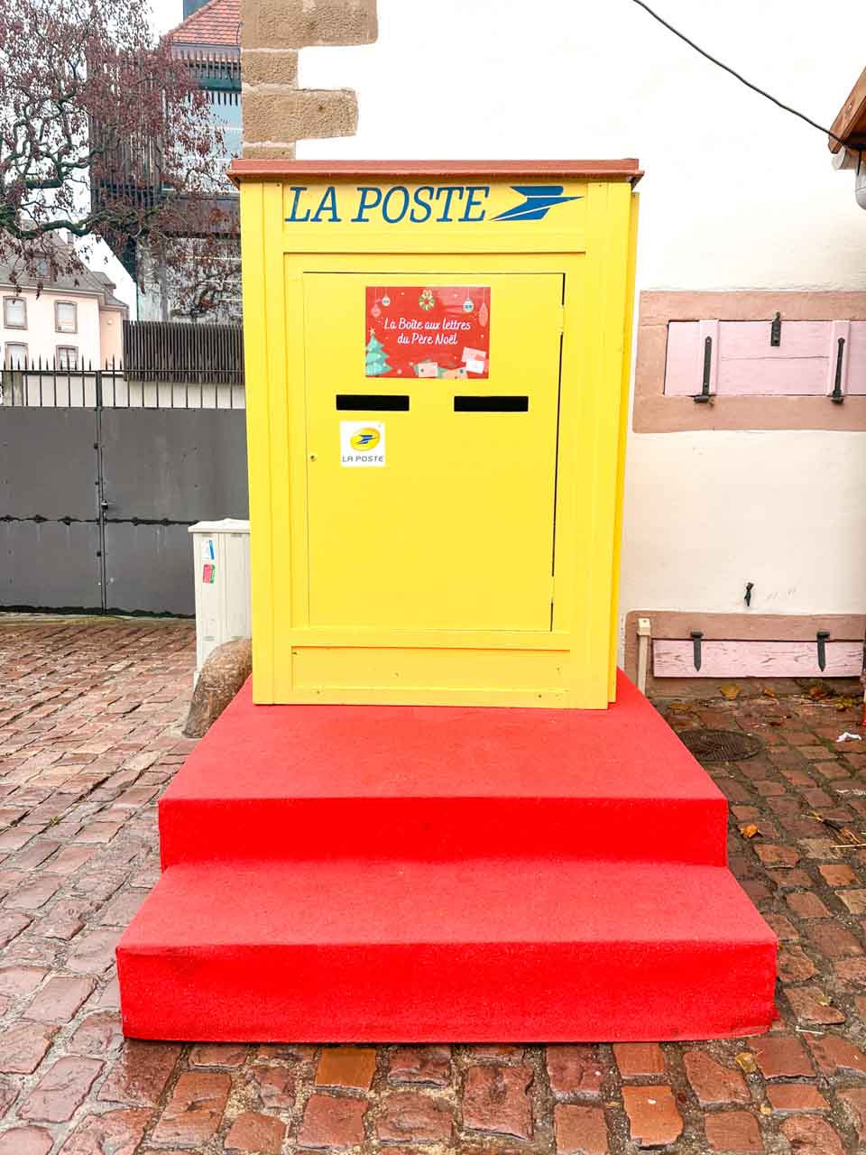 A bright yellow postbox on red steps labelled 'LA POSTE', with a festive sign for Santa's mail, stands out on a cobblestone street in Colmar