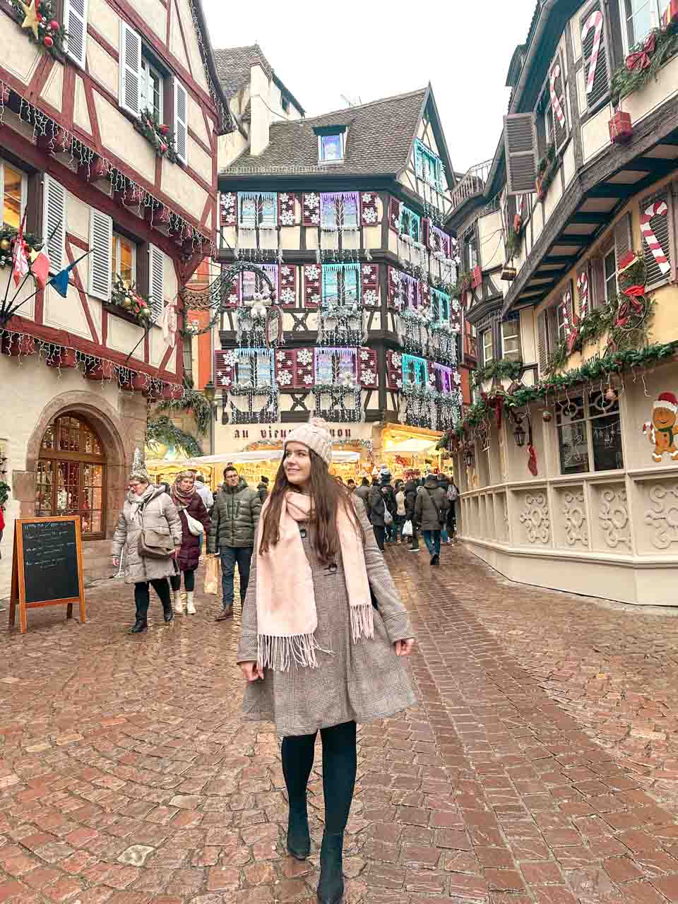 A brunette in a winter coat walking along a cobblestone street in Colmar, with beautifully decorated half-timbered houses and festive decorations in the background