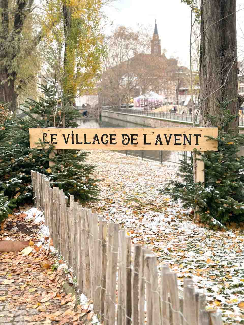 A sign that reads "Le Village de L'Avent" set against a backdrop of snow-dusted greenery, inviting visitors into a festive area on Square Louise Weiss in Strasbourg