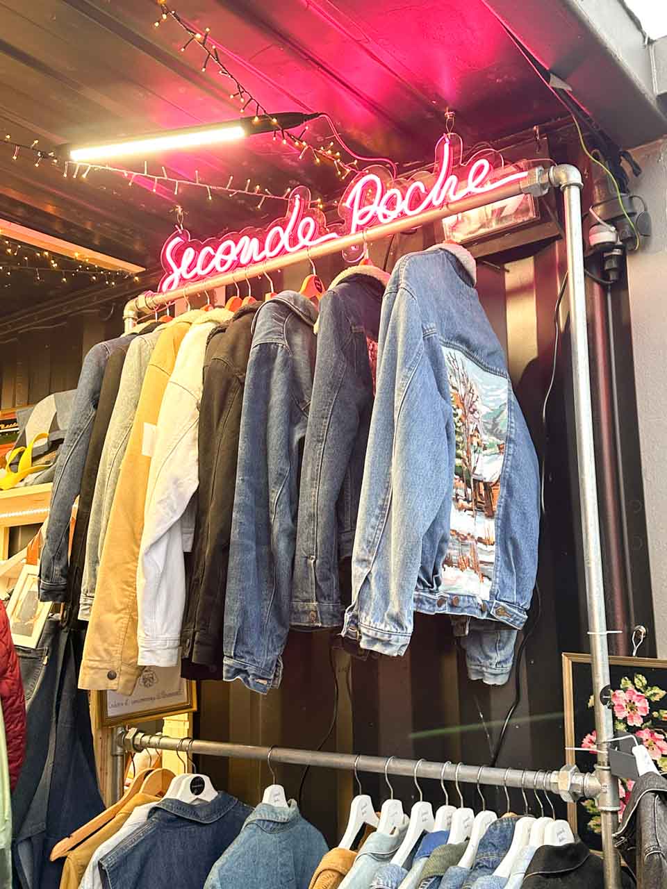 A neon sign reading "Seconde Poche" above a selection of second-hand clothing at a stall at Place Grimmeissen in Strasbourg
