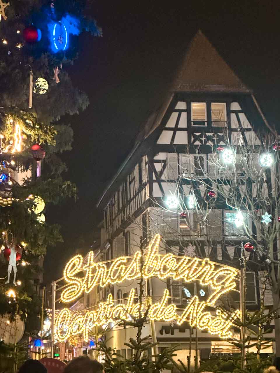 A sign that reads "Strasbourg Capitale de Noël" in bright lights with a traditional half-timbered building in the background