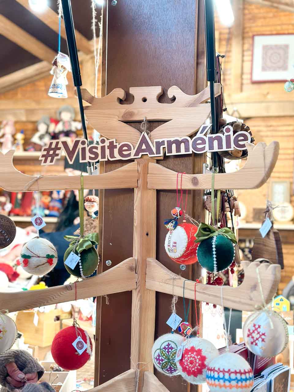 A wooden sign reading '#VisiteArmenie' pointing towards an array of handcrafted Armenian ornaments at a Christmas market stall in Strasbourg